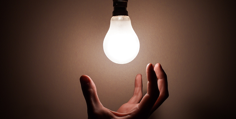 Bilden föreställer en hand som sträcker sig mot en tänd glödlampa i ett mörkt rum.